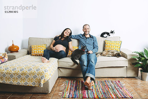 Ein glückliches Paar erwartet ein Baby  das zu Hause mit drei Katzen auf der Couch sitzt.