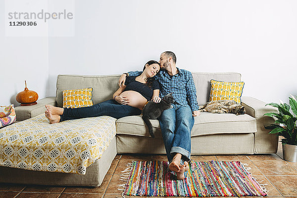 Ein glückliches Paar erwartet ein Baby  das zu Hause mit zwei Katzen auf der Couch sitzt.