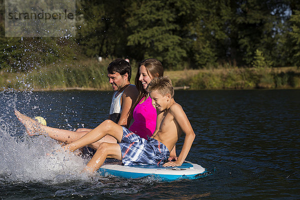 Junges Paar und ein Junge sitzen auf dem SUP Board und spritzen mit Wasser.