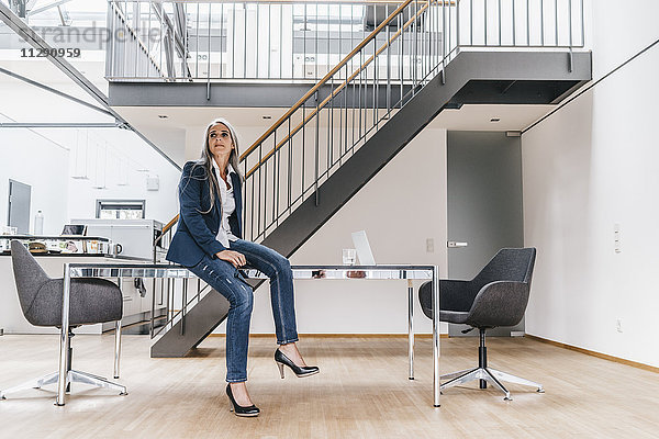 Geschäftsfrau mit langen grauen Haaren auf dem Schreibtisch im Büro sitzend