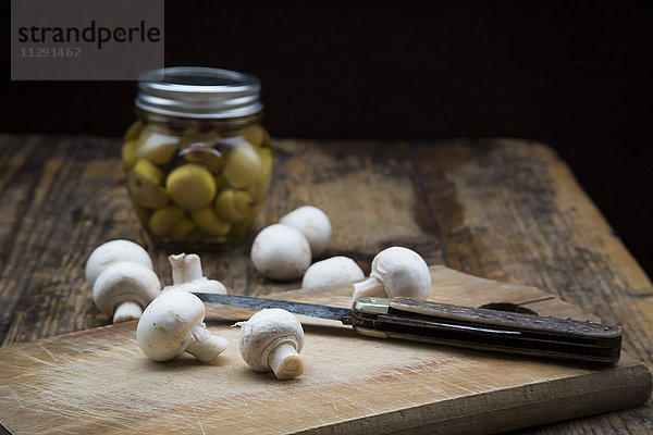 Frischer weißer Champignon und Taschenmesser auf Holzbrett und Glas eingelegter Champignons