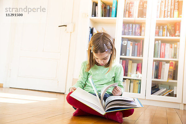 Kleines Mädchen sitzt auf dem Boden und schaut sich die Seiten eines Buches an.