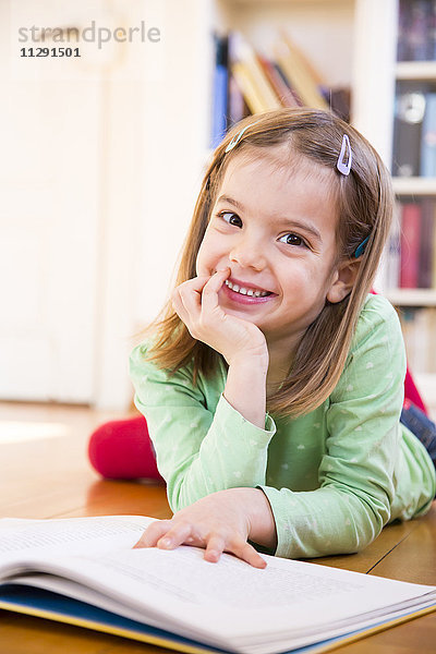 Porträt eines lächelnden kleinen Mädchens auf dem Boden liegend mit Buch