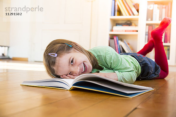 Lachendes kleines Mädchen  das mit einem Buch auf dem Boden liegt.