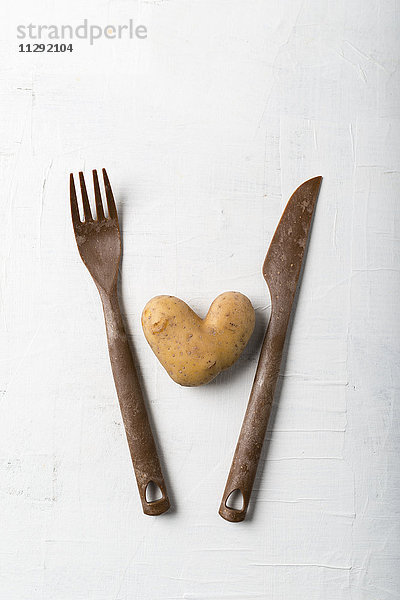 Kartoffel in Herzform zwischen Gabel und Messer auf weißem Grund