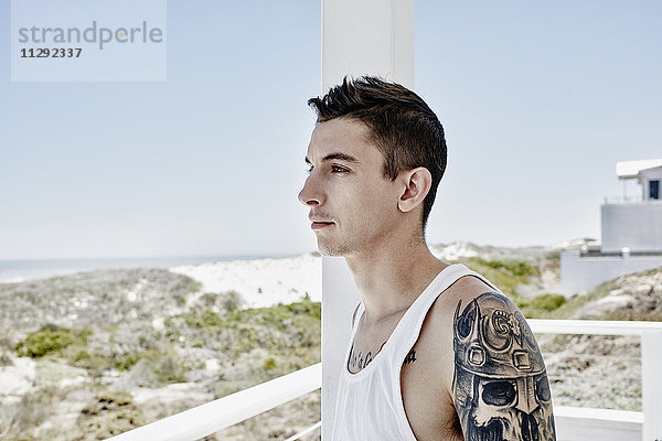 Tätowierter junger Mann auf einer Terrasse des Strandhauses mit Blick auf das Meer