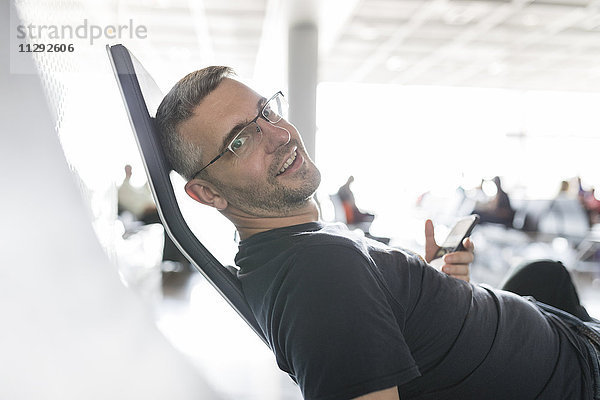 Mann sitzt im Abflugbereich des Flughafens und hält ein Smartphone.
