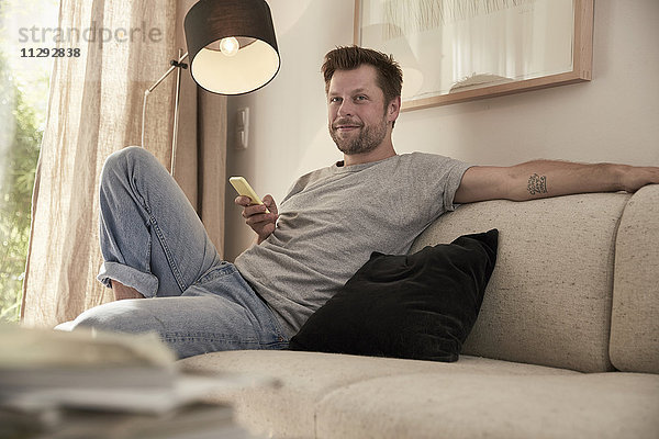 Entspannter Mann zu Hause sitzend auf der Couch mit Handy