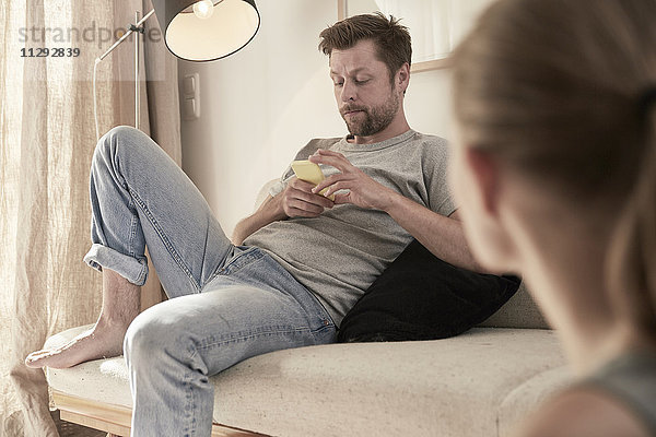 Mann zu Hause auf der Couch sitzend mit Blick auf Handy und Frauen im Vordergrund