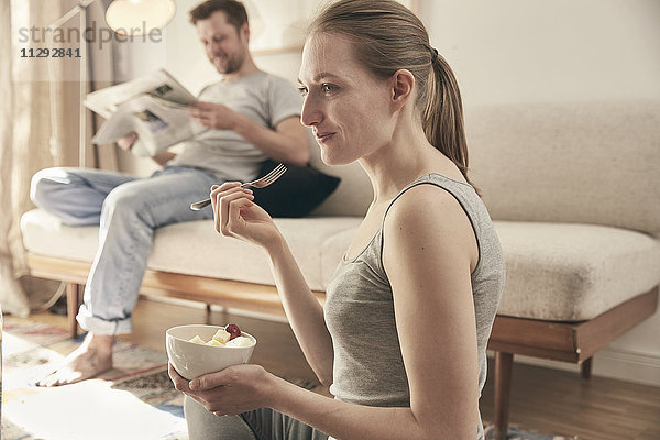 Frau isst Früchte im Wohnzimmer mit Mann im Hintergrund