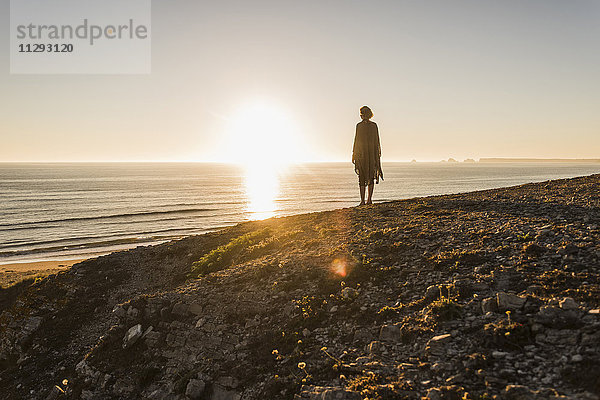 Rückansicht des jungen Mädchens  das bei Sonnenuntergang auf dem Hügel steht und auf das Meer schaut.