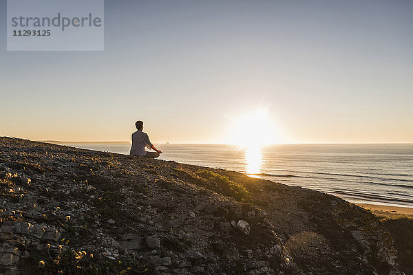 Rückansicht einer Frau  die bei Sonnenuntergang auf einer Klippe meditiert.