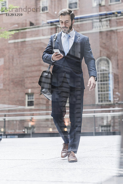 USA  New York City  Geschäftsmann  der in Manhattan spazieren geht und sich das Handy ansieht.