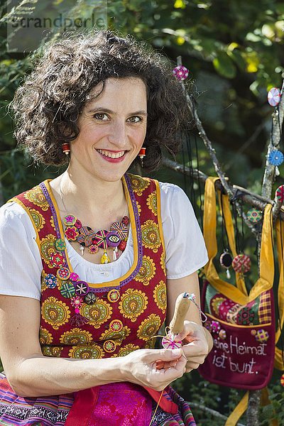 Knopfmacherin  junge Frau in buntem Dirndl mit Posamentenknopf Collier  sitzt im Garten und arbeitet an Posamentenknopf  Ichenhausen  Bayern  Deutschland  Europa