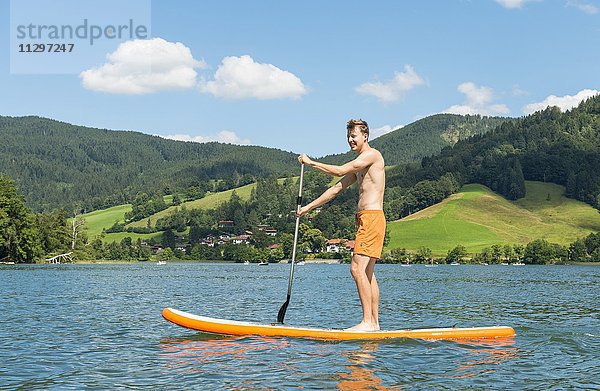 Junger Mann auf einem Stehpaddel  Stand-Up-Paddel oder SUP auf einem See  Schliersee  Oberbayern  Bayern  Deutschland  Europa
