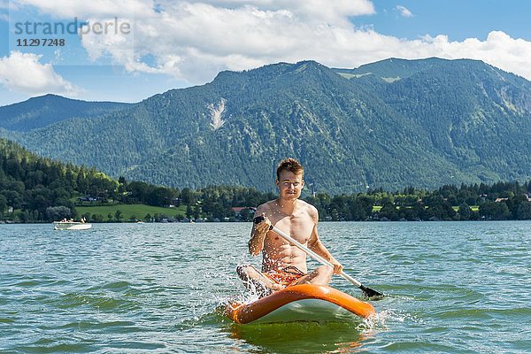 Junger Mann sitzt auf einem Board  Stand-Up-Paddel oder SUP auf einem See  Schliersee  Oberbayern  Bayern  Deutschland  Europa