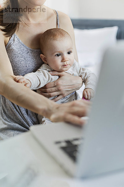 Mutter hält Baby-Tochter und arbeitet am Laptop