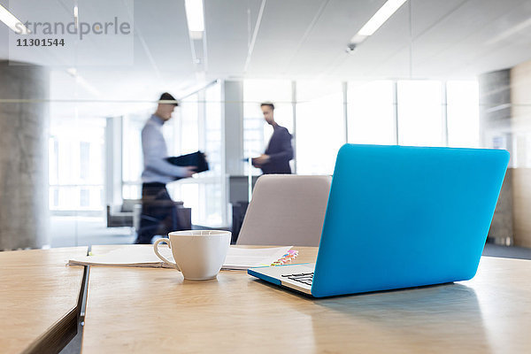 Laptop und Kaffeetasse auf Konferenzraumtisch mit Geschäftsleuten im Hintergrund