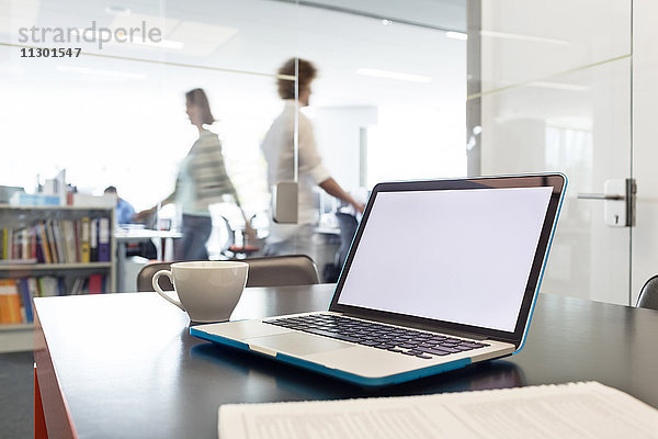 Laptop und Kaffeetasse auf dem Schreibtisch mit Geschäftsleuten im Hintergrund
