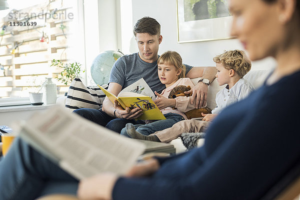 Vater liest Geschichtenbuch für Kinder mit Mutter im Wohnzimmer