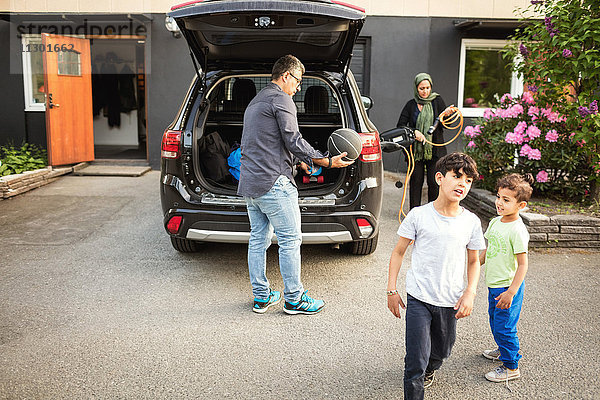Jungen stehen  während Vater und Mutter vor dem Haus am Auto stehen.