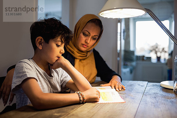 Mutter und Sohn lesen Buch unter der beleuchteten Schreibtischlampe zu Hause