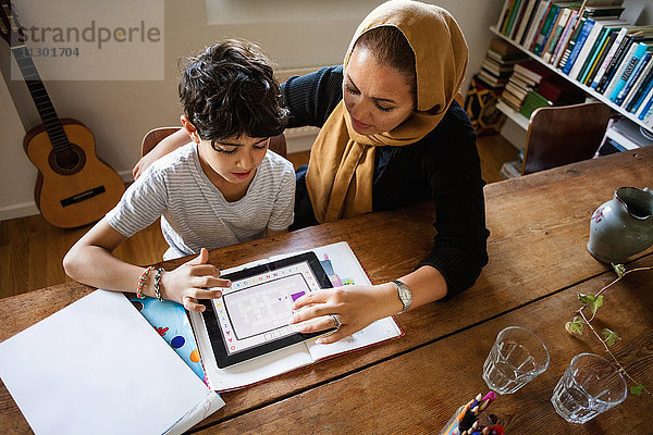 Hochwinkelansicht der Mutter  die den Sohn bei der Verwendung des digitalen Tabletts während des Studiums zu Hause unterstützt.