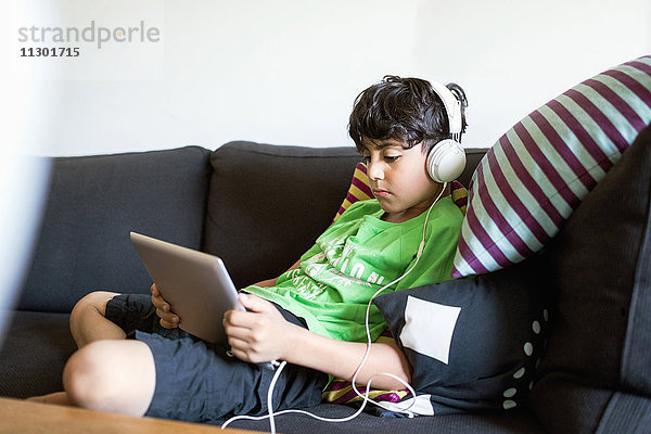 Junge  der Musik über ein digitales Tablett hört  während er zu Hause auf dem Sofa sitzt.
