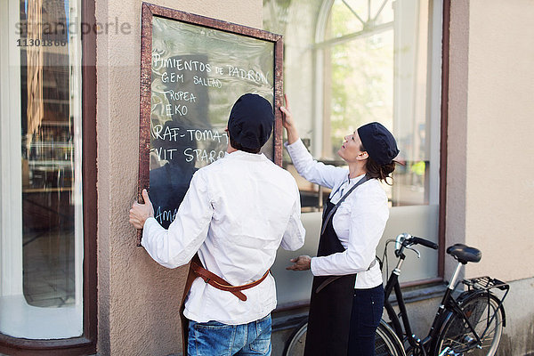 Männliche und weibliche Besitzer  die eine Tafel an der Wand außerhalb des Lebensmittelladens anbringen.