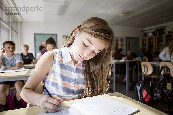 Mädchen beim Schreiben im Buch am Schreibtisch mit Schülern im Hintergrund