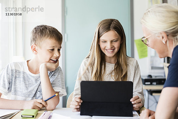 Fröhliches Mädchen mit digitalem Tablett  während Lehrer und Junge in der Schule im Klassenzimmer zuschauen.