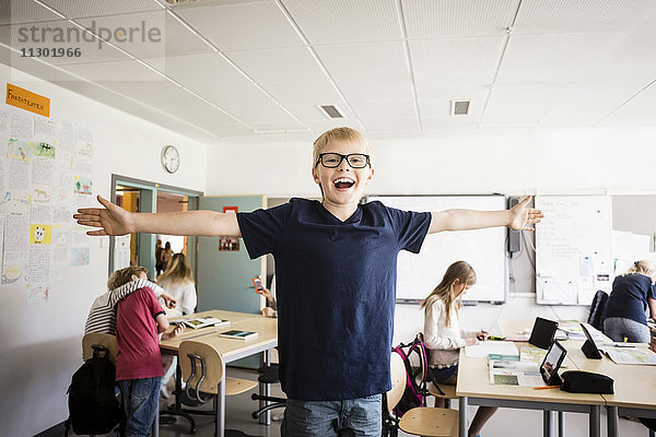 Glücklicher Junge steht mit ausgestreckten Armen gegen Freunde im Klassenzimmer.