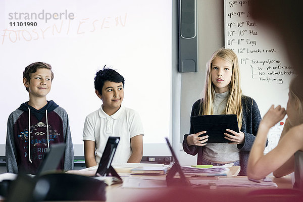 Fröhliches Mädchen beim Präsentieren mit digitalem Tablett inmitten von Schülern im Klassenzimmer