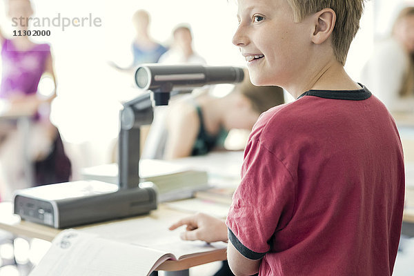 Fröhlicher Junge liest Buch unter der Kamera auf dem Schreibtisch im Klassenzimmer