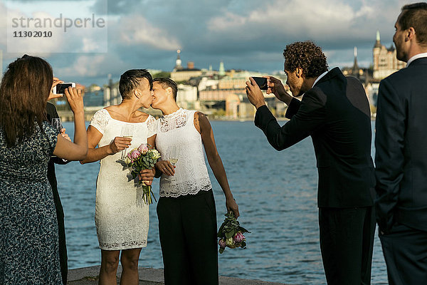 Glücklicher Mann mit Freunden beim Fotografieren eines lesbischen Paares  das sich auf der Hochzeit küsst.