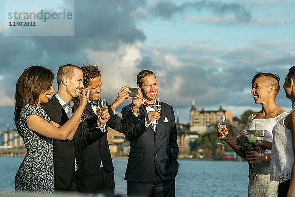 Glücklicher Mann mit Freunden beim Fotografieren eines lesbischen Paares bei der Hochzeit