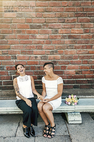 Fröhliches jungvermähltes lesbisches Paar auf der Bank an der Wand sitzend