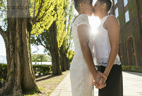 Seitenansicht eines frisch verheirateten lesbischen Paares  das sich küsst  während es sich auf dem Fußweg an den Händen hält.