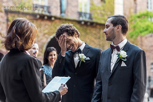 Schwuler Mann sieht schüchternen Partner an  während er während der Hochzeitszeremonie vor dem Priester steht.
