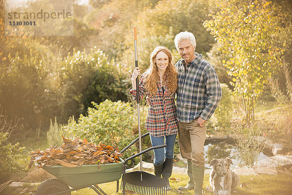 Portrait lächelndes Paar mit Hund bei der Gartenarbeit Laubharken im Herbstgarten