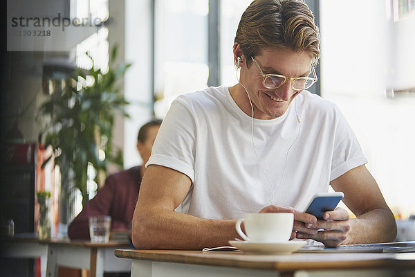 Lächelnder Mann mit Kopfhörern  der ein Mobiltelefon benutzt und Kaffee in einem Café trinkt