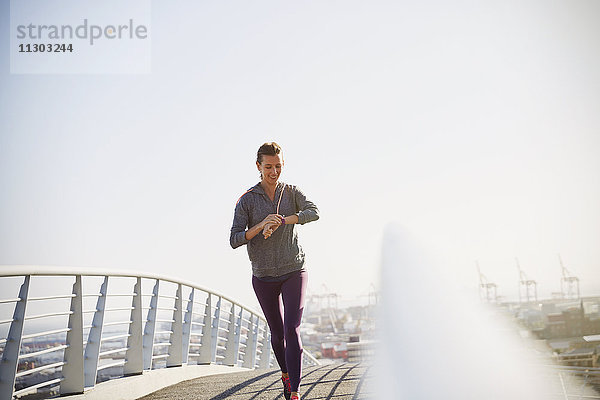 Lächelnde Läuferin  die auf einer sonnigen Fußgängerbrücke ihren Smartwatch-Fitness-Tracker überprüft