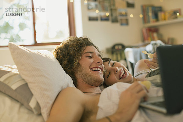 Lächelndes junges Paar liegt im Bett und benutzt einen Laptop