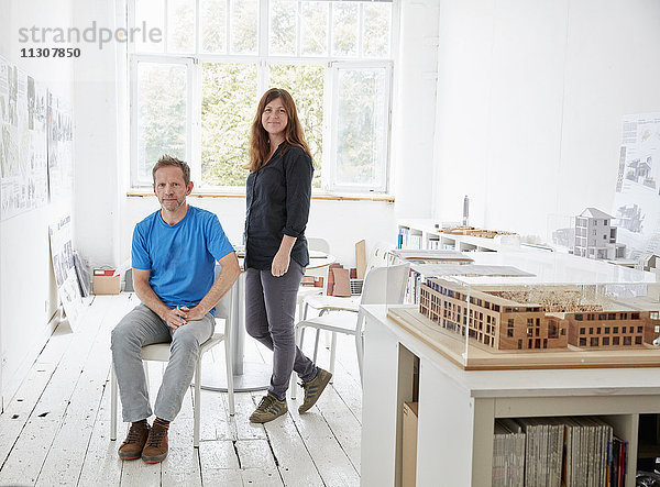 Ein modernes Büro mit weißen Wänden und Böden. Ein sitzender Mann und eine stehende Frau. Ein Architekturmodell auf einem Tisch.