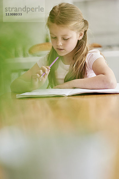 Ein Mädchen  das mit einem Bleistift an einem Tisch sitzt und seine Hausaufgaben macht.