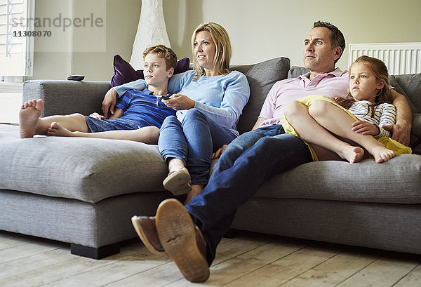 Eine vierköpfige Familie mit Eltern und einem Mädchen und einem Jungen  die zusammen auf dem Sofa sitzen und fernsehen.