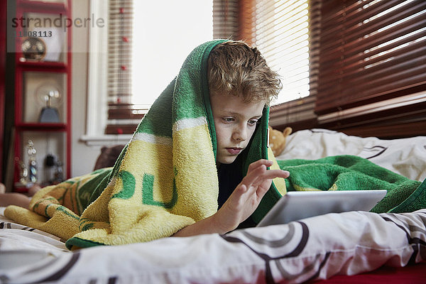 Ein Junge mit einem Handtuch über dem Kopf  der auf seinem Bett liegt und ein digitales Tablett benutzt.