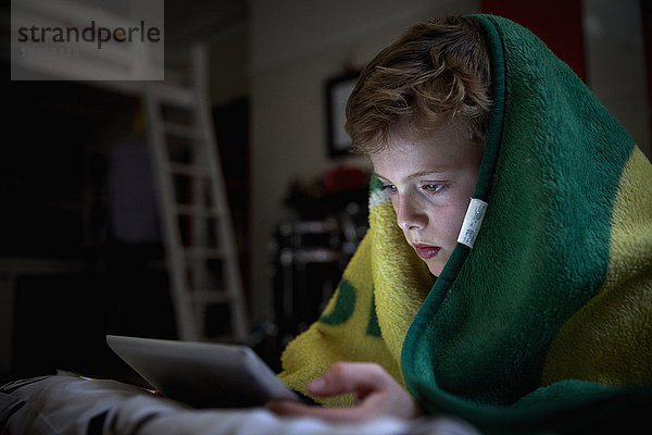 Ein Junge mit einem Handtuch über dem Kopf  der auf seinem Bett liegt und ein digitales Tablett benutzt.