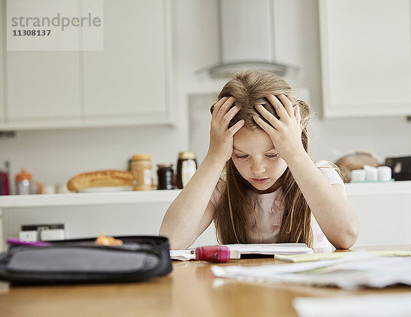 Ein Mädchen sitzt an einem Tisch mit den Händen auf dem Kopf und schaut auf ihre Hausaufgaben.