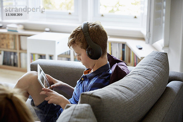 Ein Familienhaus. Ein Junge auf einem Sofa  der ein digitales Tablet benutzt und Kopfhörer trägt.